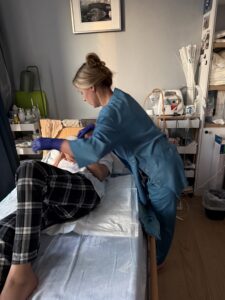 Zdjęcie. Kobieta w jednorazowym fartuchu medycznym pochyla się nad pacjentem leżącym w łóżku